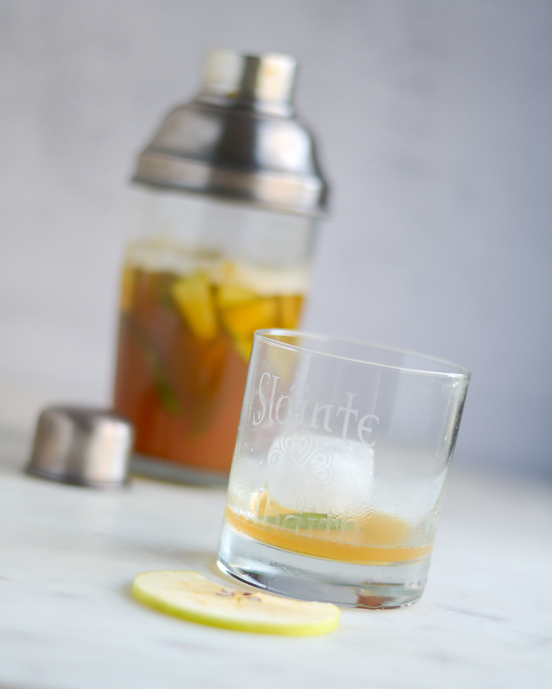 empty glass of mullen tea drink in a low ball glass with apple wedge garnish and cocktail shaker in the background