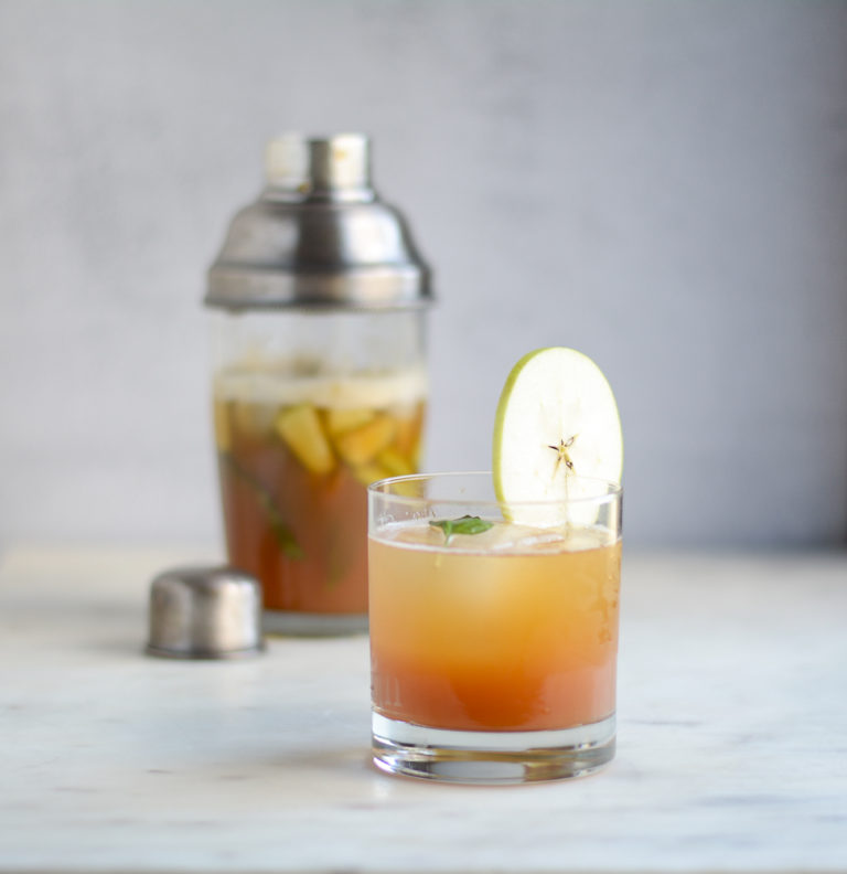 square shot of mullen tea drink in a low ball glass with apple wedge garnish and cocktail shaker in the background