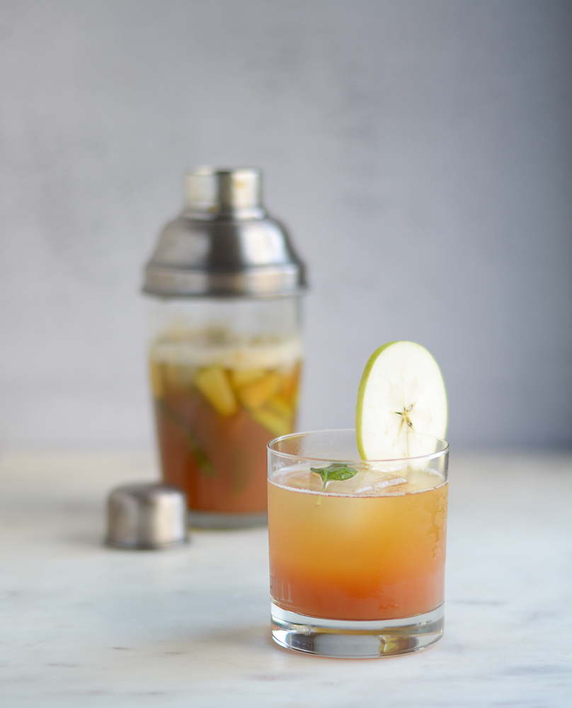 vertical shot of mullen tea drink in a low ball glass with apple wedge garnish and cocktail shaker in the background