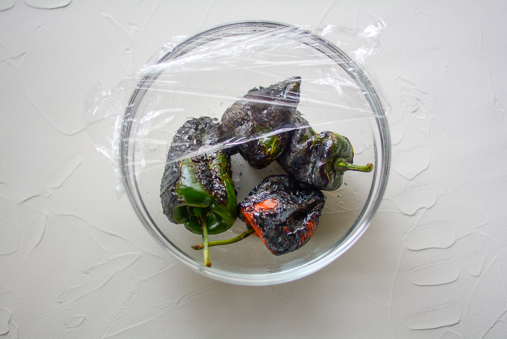 Flame roasted chiles in a bowl with plastic wrap